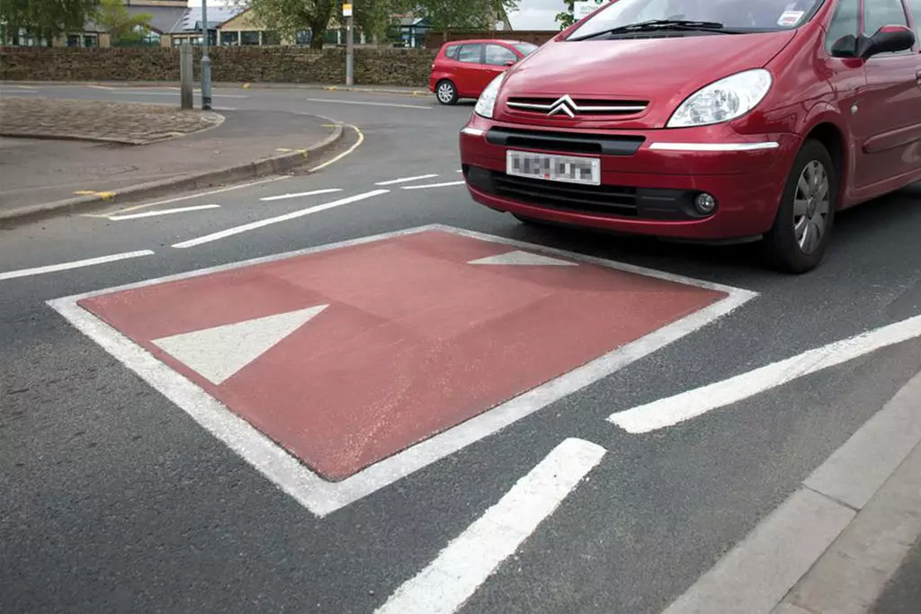 A red car is about to drive over a red concrete speed cushion with white markings.