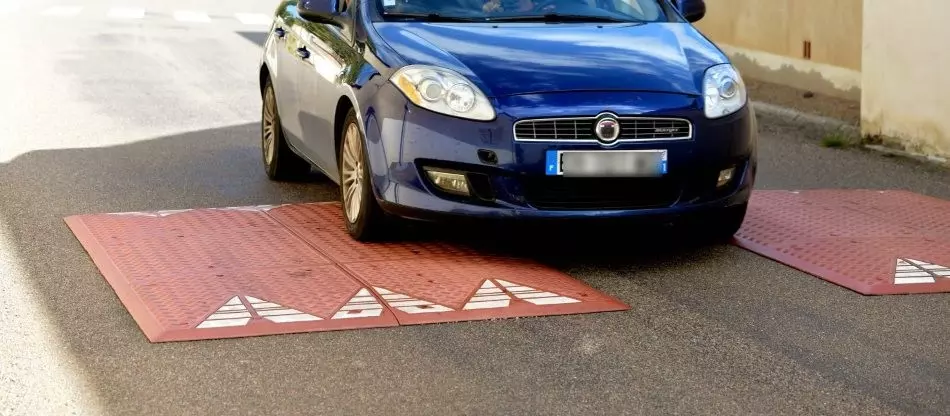 A navy blue car is driving over two red speed cushions made of rubber without noise.