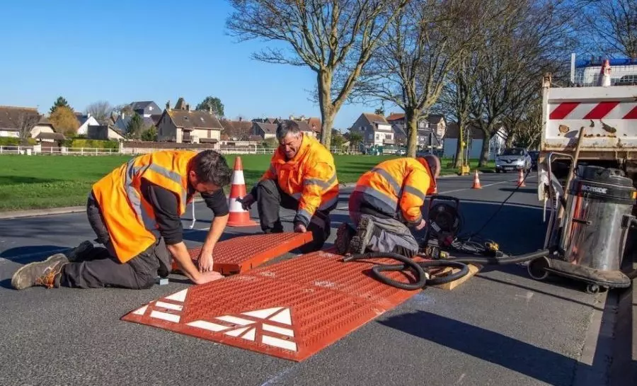 Three men in orange are installing a red rubber speed cushion.